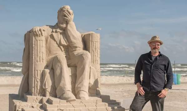 Sand sculpture of Lincoln with his hand to his face and cracks in the sand. The sand sculptor stands close to his work, smiling.