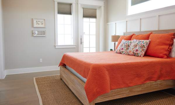 A bed with a wooden headboard and orange quilt and floral pillows sits in the middle of a bright, light bedroom.
