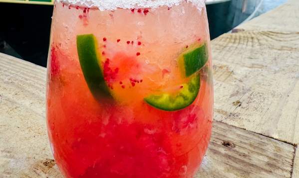 A red strawberry margarita with floating chunks of jalapeno and a salted rim in a stemless wine glass sits on the edge of a porch in front of a pirate ship in the water.