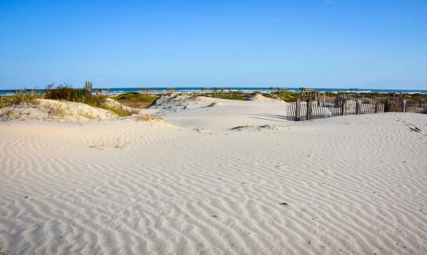 Groomed sand with grassy dunes in the background. Water is visible in the background.