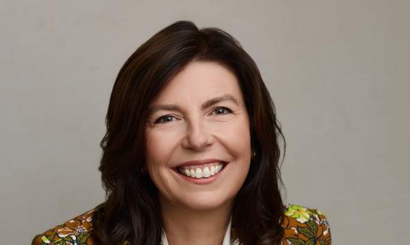 Headshot of Leslie Bruce. She's sitting, smiling at the camera with her hands crossed. She's wearing a white blouse and a floral-print blazer.