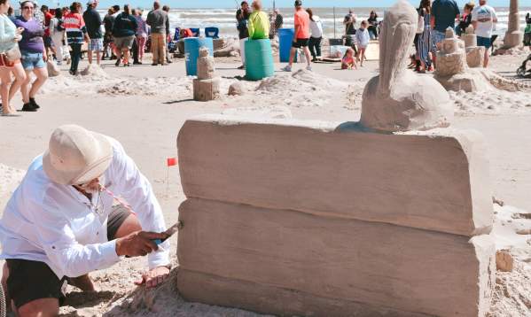 A man crouches next to a block of sand and carves it. A rudimentary pelican is carved on top of the block of sand.