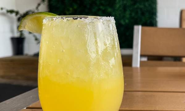 A yellow margarita in a stemless glass sits on a table. In the background is a greenery wall with a neon sign that says "I <3 Tortuga's"