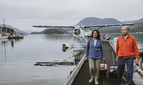 Two people wheel their suitcases along the dock after disembarking from a floatplane.