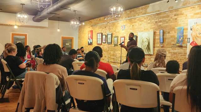 In Fayetteville, NC, Audience seated in folding chairs listening to a poet at a microphone in an art gallery with paintings displayed on a brick wall.