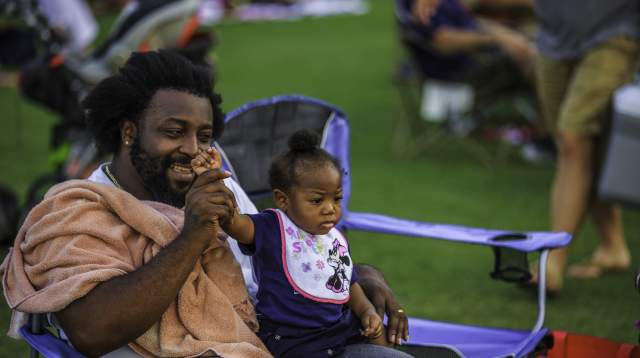 Father and child sitting together at Festival Park during a Fourth of July celebration, watching the event with other attendees nearby.