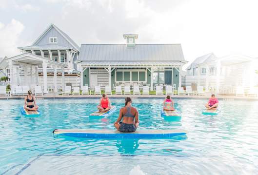 A woman with crossed legs sits atop a surf board floating in a pool, back to the camera. Four women, also with crossed legs, sit atop surf boards facing the instructor.