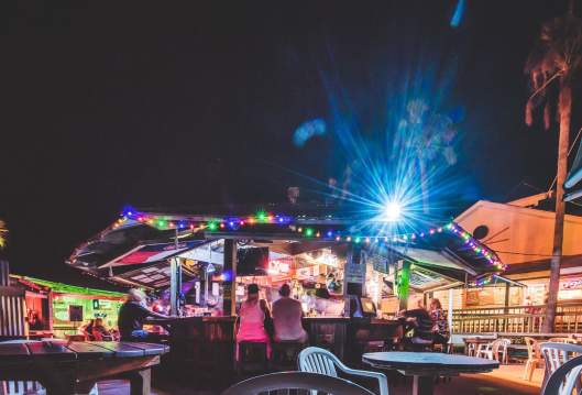 Small groups of people sit at an outdoor bar at night. The bar is decorated with bright, multicolored lights. Tables and chairs are visible in the foreground.
