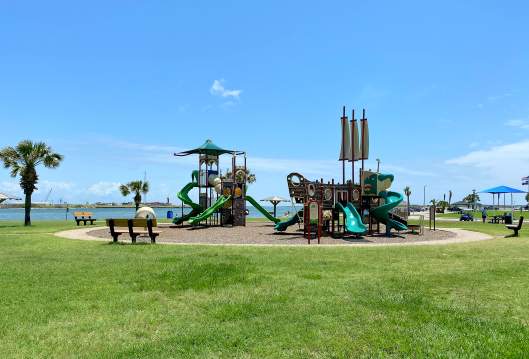 A round gravel area surrounded by lush green grass holds a large playground. The playground is green and brown with slides, ship sails, and more. Water is in the background.