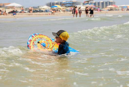 A child in a ball cap swims with a tie-dye boogie board near the beach