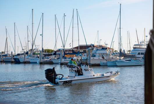 A fishing boat with two people in the driver's seat putters through a marina with rows of sailboats behind