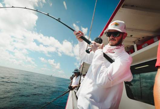 A guy in a white shirt, sunglasses, and a hat casts a line off the side of a boat in the deep sea