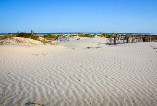 Groomed sand with grassy dunes in the background. Water is visible in the background.