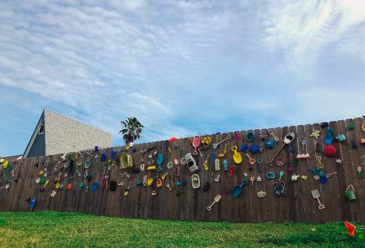 A fence is dotted with colorful discarded beach toys—shovels, cars, buckets, and more. A sky covered in white clouds is above and a house is visible in the left background