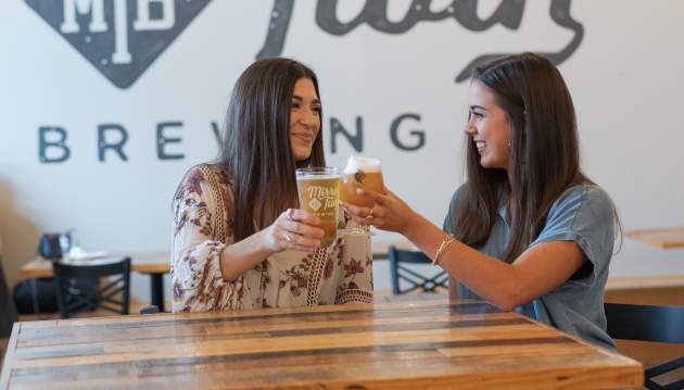 Two women cheers their beers and smile inside the tasting room of Mirror Twin Brewing.