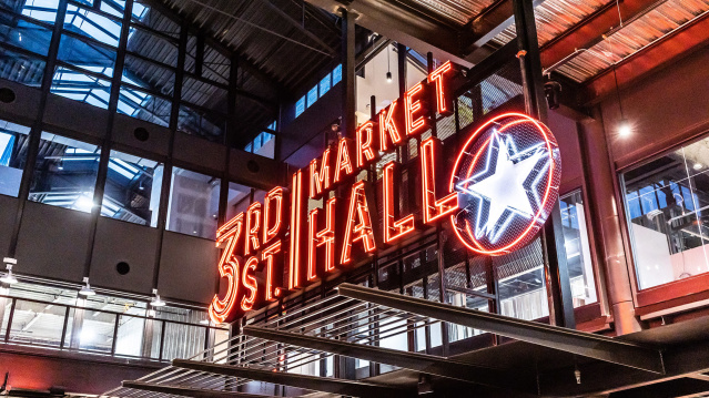 Neon sign reading ‘3rd Street Market Hall’ with a glowing star, inside a modern industrial-style building with glass walls and metal beams.