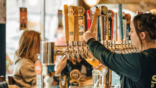 A bartender pours a draft beer from a row of colorful tap handles while customers sit at the bar in a cozy pub setting.