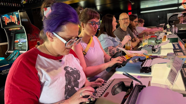 A group of people sit side by side at a bar counter, each using a typewriter. The person in the foreground has purple hair tied back with a scrunchie, glasses, and a red-and-white shirt with a graphic print. Others focus intently on their typewriters, with drinks and cans on the counter. Behind them, arcade machines, merchandise, and a TV screen are visible, creating a lively retro atmosphere.