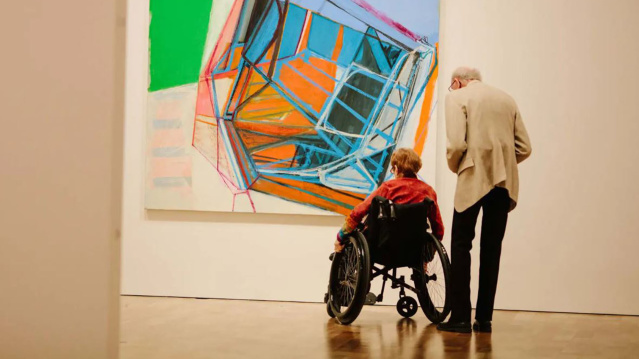 Two elderly people, one in a wheelchair, admire a large, colorful abstract painting in a bright art gallery.