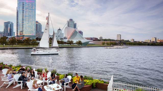 a group of people eating on the patio in front of the Milwaukee skyline with a sailboat in the harbor