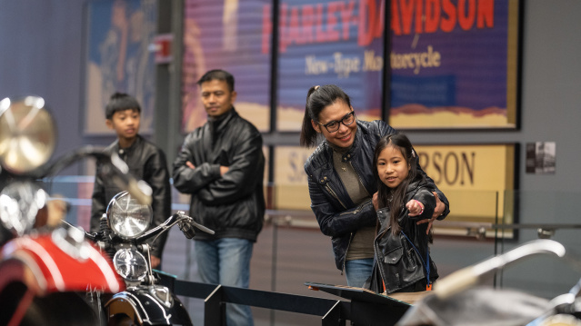 A family of four, all dressed in black leather jackets, visits the Harley-Davidson Museum. A smiling woman and a young girl in the foreground point at a vintage motorcycle display, while two other family members stand behind them observing.