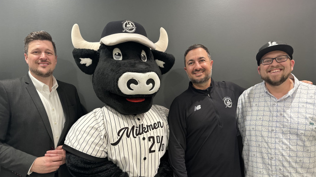 Three men pose for a photo with the Milwaukee Milkmen mascot, a smiling black and white cow wearing a pinstriped baseball uniform and cap, standing in front of a plain dark gray wall.