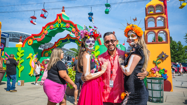 Three people pose and smile at a vibrant outdoor festival in Milwaukee. Two wear colorful Day of the Dead-style face paint and flower crowns, standing next to a man in sunglasses. Behind them is a festive archway and colorful decorations, with people enjoying the event under a bright blue sky.