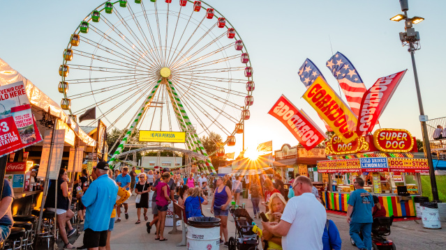 A colorful summer fair scene at sunset featuring a large Ferris wheel with green, yellow, red, and orange gondolas in the background. The foreground is bustling with people walking, eating, and enjoying the fair. Brightly lit food stands advertise corn dogs, lemonade, and "Super Dogs," with American flags waving above. A yellow sign at the Ferris wheel entrance reads "$6 PER PERSON." The sun sets behind the attractions, casting a warm glow over the festive atmosphere.