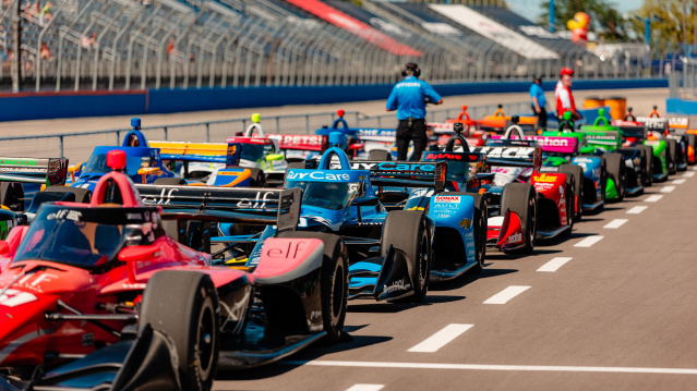 IndyCar race cars lined up on the track at the Milwaukee Mile with drivers and officials preparing for the start of the race, as spectators watch from the grandstands under a sunny sky.