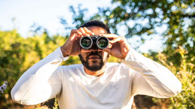 A man in a white long-sleeved shirt holds binoculars up to his eyes while standing outdoors in a sunny, tree-filled setting with green foliage and dappled sunlight in the background.