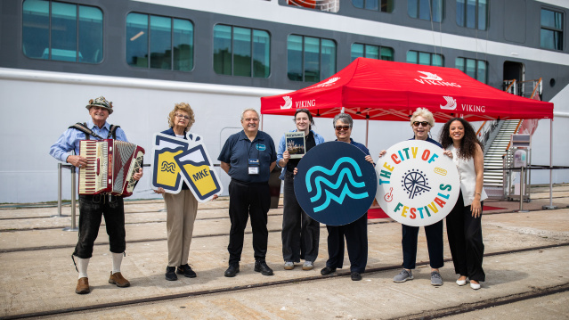 A welcoming committee with an accordion player stands before a cruise ship, holding Milwaukee-themed signs including an MKE logo and "City of Festivals" circular display, under a red Viking Cruises tent.