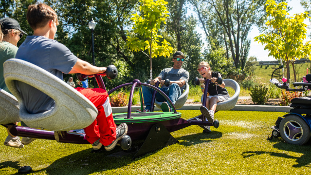 Children of varying abilities play together on an accessible playground spinner, with one child using a power wheelchair alongside others seated on the equipment, surrounded by trees and greenery on a sunny day.