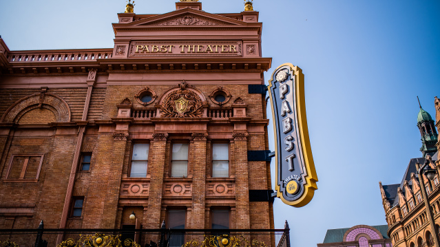 The exterior of the historic Pabst Theater in Milwaukee, featuring its ornate brick façade, gold architectural details, and large vertical “Pabst Theater” marquee sign against a clear blue sky.