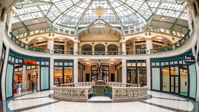Interior of an ornate shopping arcade with a large glass-domed ceiling, decorative railings and storefronts surrounding a central atrium