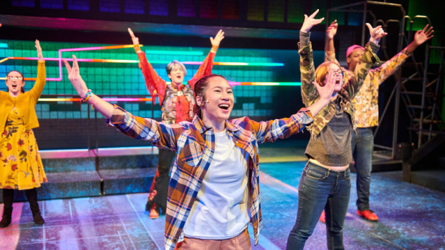 A group of young performers on a brightly lit stage throw their arms up mid-dance, smiling and full of energy, with colorful set pieces glowing behind them.