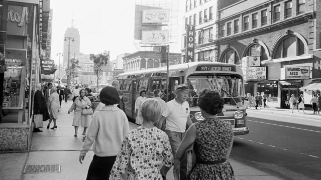 A historic black-and-white photograph shows a busy downtown Milwaukee street lined with storefronts, pedestrians, and a city bus traveling along the road. Vintage signage, mid-century architecture, and everyday street activity capture the character of Milwaukee's urban core in an earlier era.