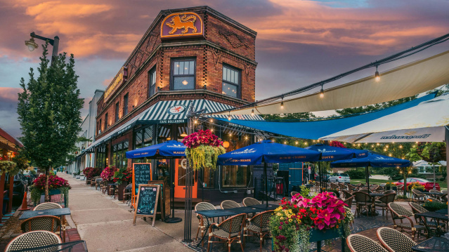 A brick building set against a sky at dusk, with orange and blue tones, a patio lit up with string lights, blue umbrellas and pink and yellow flowers.