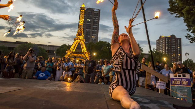 A fire performer in a striped costume kneels and raises flaming batons during a lively Bastille Days event in Milwaukee, with a crowd watching and a glowing Eiffel Tower replica lit up in the background.