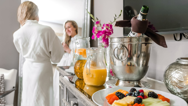 Guest in a white robe enjoys a relaxing hotel morning with fresh fruit, orange juice, and champagne on a tray beside an orchid and elegant décor.