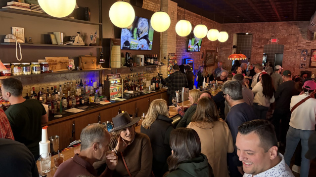 A crowded bar with warm lighting and exposed brick walls. People are gathered closely together, talking and drinking. A long bar counter is lined with bottles and glassware, and several round hanging lights illuminate the space. Two TVs mounted on the back wall display a show, and the overall atmosphere appears lively and social.