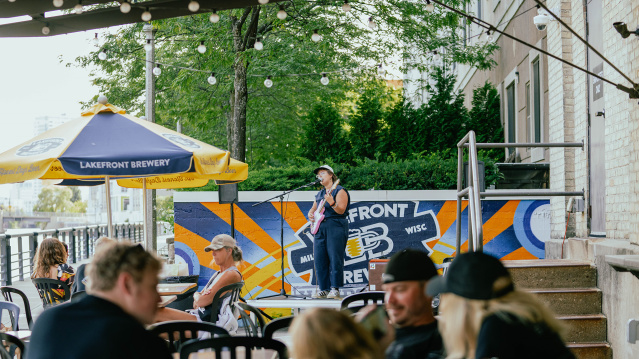 A woman in a navy blue romper singing into a microphone on a stage, holding a pink guitar, with people in front of her sitting at tables under gold and blue umbrellas on a patio along the riverfront with trees in the background.