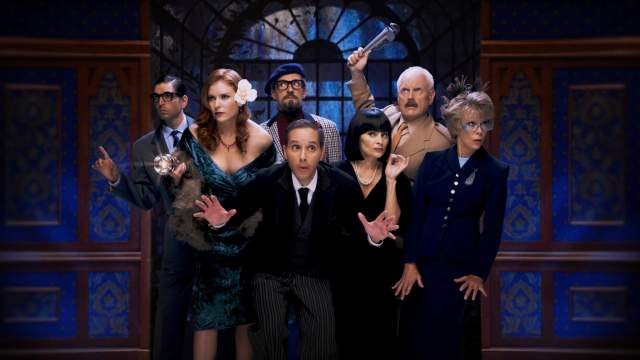 Seven Clue characters pose together in a dramatic mansion setting, wearing colorful, vintage-style costumes and reacting with surprise and suspicion, including one man holding a raised wrench
