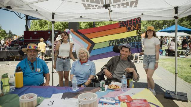 Two people standing holding a pride flag that says "Every one is welcome here". Three people sit in front of the flag.