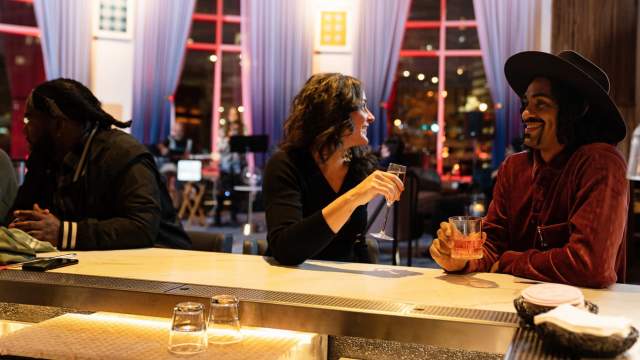 Two people sit at a stylish bar engaged in a lively conversation, each holding a drink. The woman smiles while talking to the man in a wide-brimmed hat. The setting is warmly lit with modern decor, including large windows, blue curtains, and ambient city lights in the background. Another person sits further down the bar, looking away.