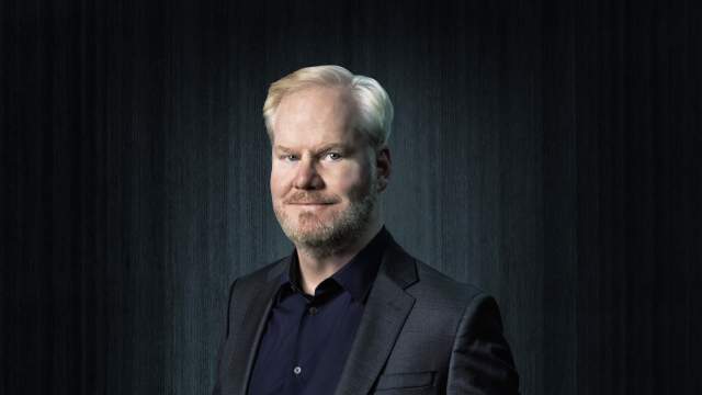 Jim Gaffigan in a suit in front of a black curtain