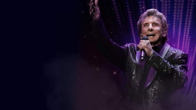 Barry Manilow singing into a microphone with his opposite hand extended out