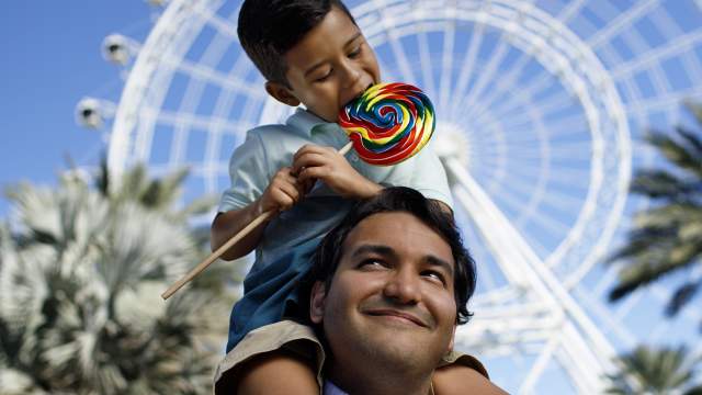 A boy on top of his dad's shoulders and eating a lollipop in front of The Wheel at ICON Park