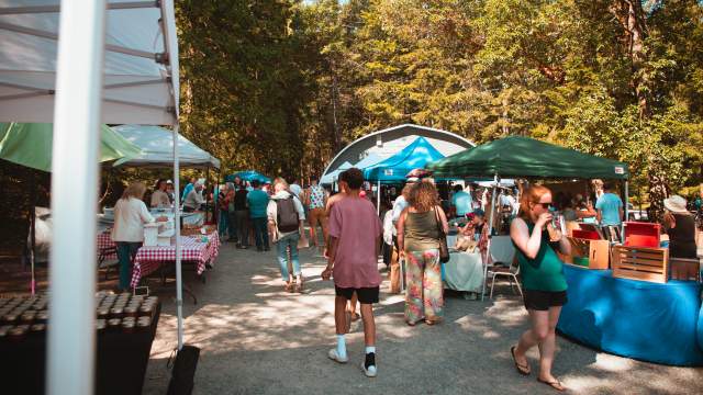 Visitors wander between vendor stalls at a busy outdoor market on Gabriola Island, set among tall trees.