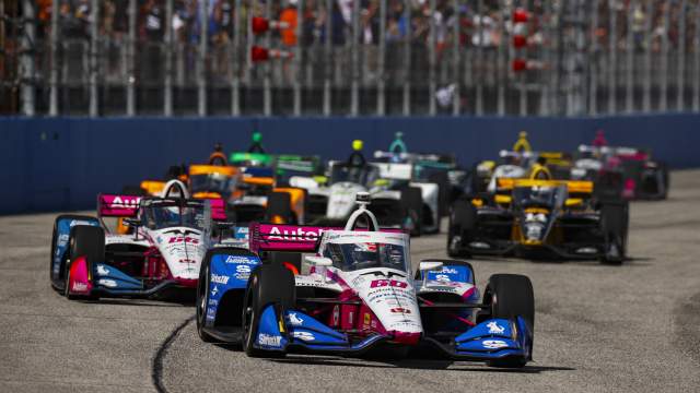 A dynamic shot of several IndyCars speeding along a track, led by a white, pink, and blue #60 car, with a crowd visible in the background.