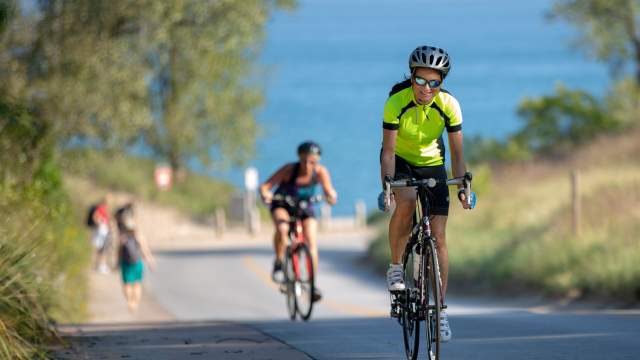 Bicyclists ride up hill on a paved road. A lake is in view in the background.